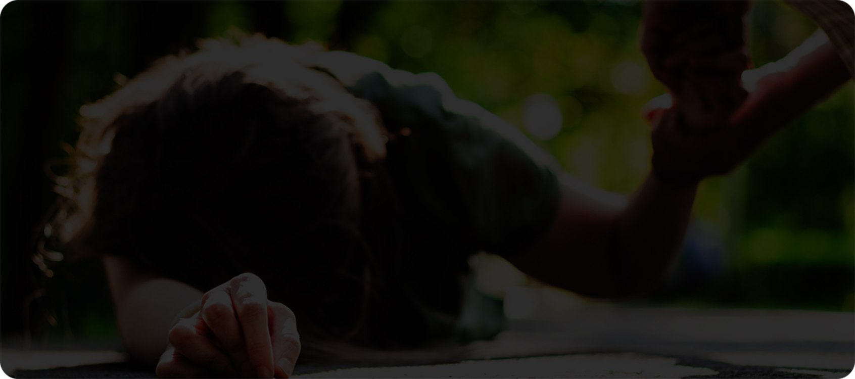 femme couchée face contre terre, s’appuyant sur son avant-bras droit; une personne près d’elle tient sa main gauche et semble l’aider à se relever